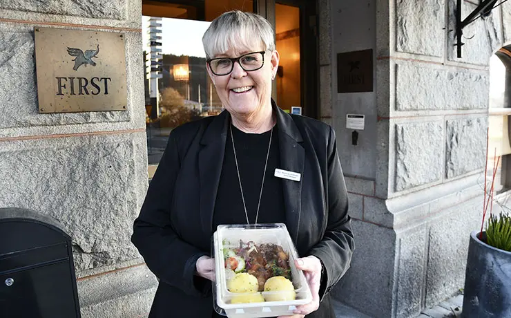 En kvinna i svart kostym står framför en hotellentré och håller fram en matlåda med genomskinligt lock i handen.