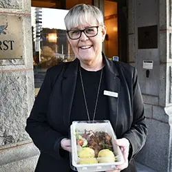 En kvinna i svart kostym står framför en hotellentré och håller fram en matlåda med genomskinligt lock i handen.