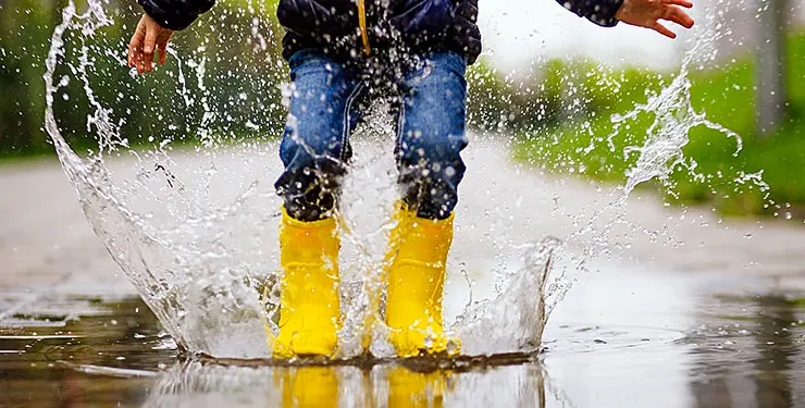 Ett barn i gula stövlar hoppar i en stor vattenpöl så att det stänker