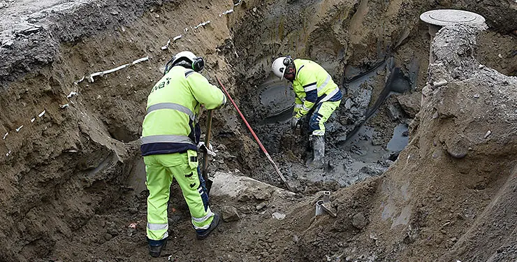 Två män i varselkläder gräver fram ett rör i en stor grop