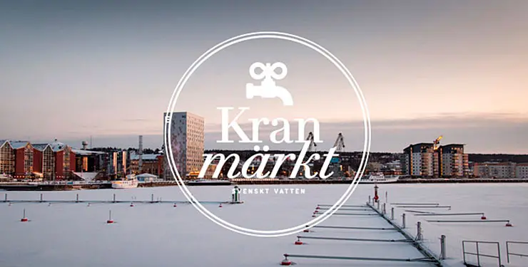 Logotype för Svenskt Vattens hållbarhetsmärkning Kranmärkt med vy av Örnsköldsviks vinterklädda inre hamn i bakgrunden