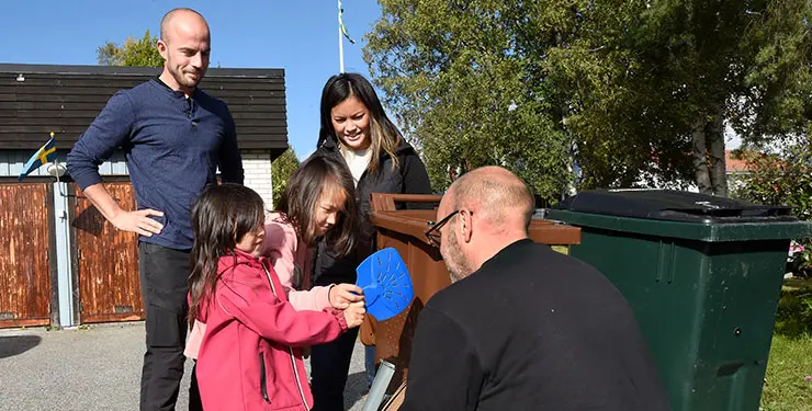 En familj står vid ett brunt sopkärl, barnen håller i en blå slaskskrapa