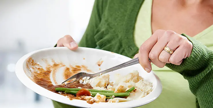En kvinna i grön tröja skrapar matrester från en vit tallrik med en gaffel