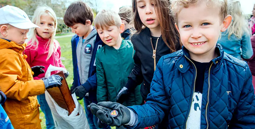 En pojke håller upp en kapsyl i handen, många barn i bakgrunden tittar nyfiket på