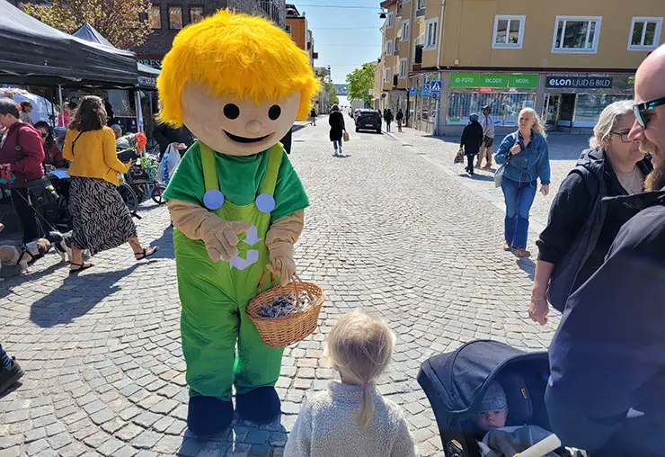 Figuren Mivo i stort gult hår och gröna kläder med en korg i handen står på ett torg och vinkar till ett litet barn.