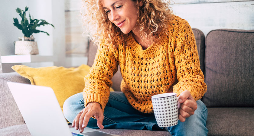 En kvinna i stort lockigt hår sitter i en soffa och dricks te och tittar på en laptop