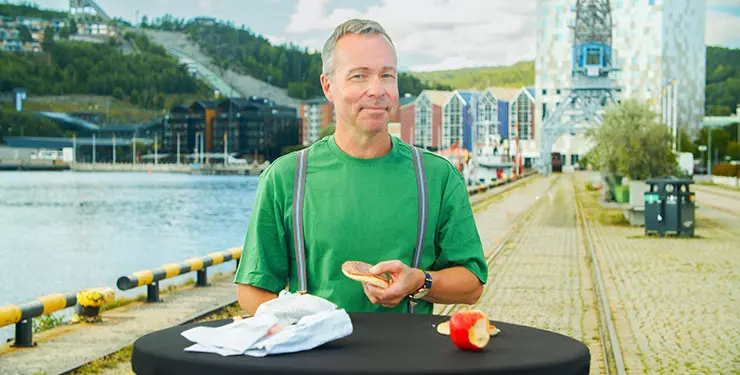 Man i grön t-shirt och hängslen som håller i en smörgås vid vattnet i Örnsköldsviks hamn