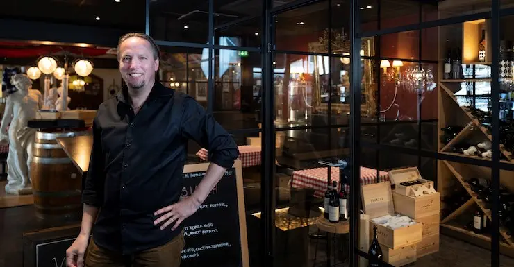 En glad man i svart skjorta ler mot kameran och interiören från en restaurang i bakgrunden